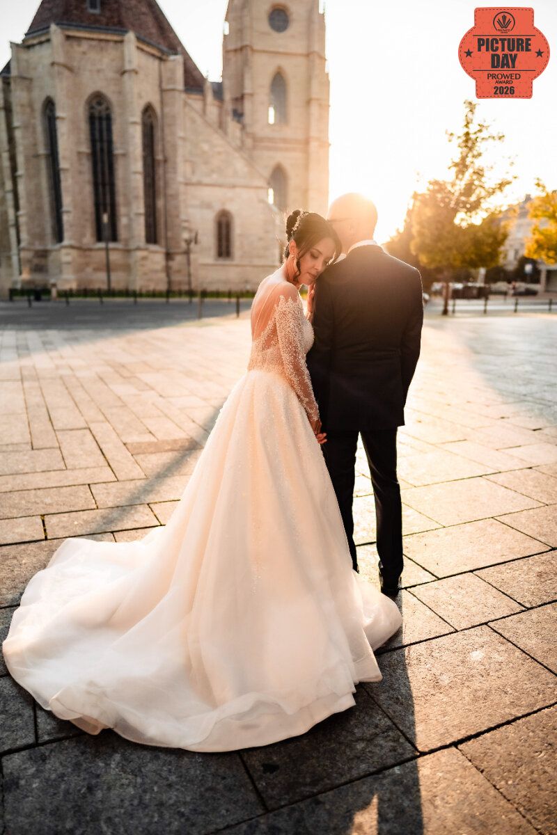 Bride and groom embrace at sunset in front of historic church, her lace gown trailing on cobblestones.