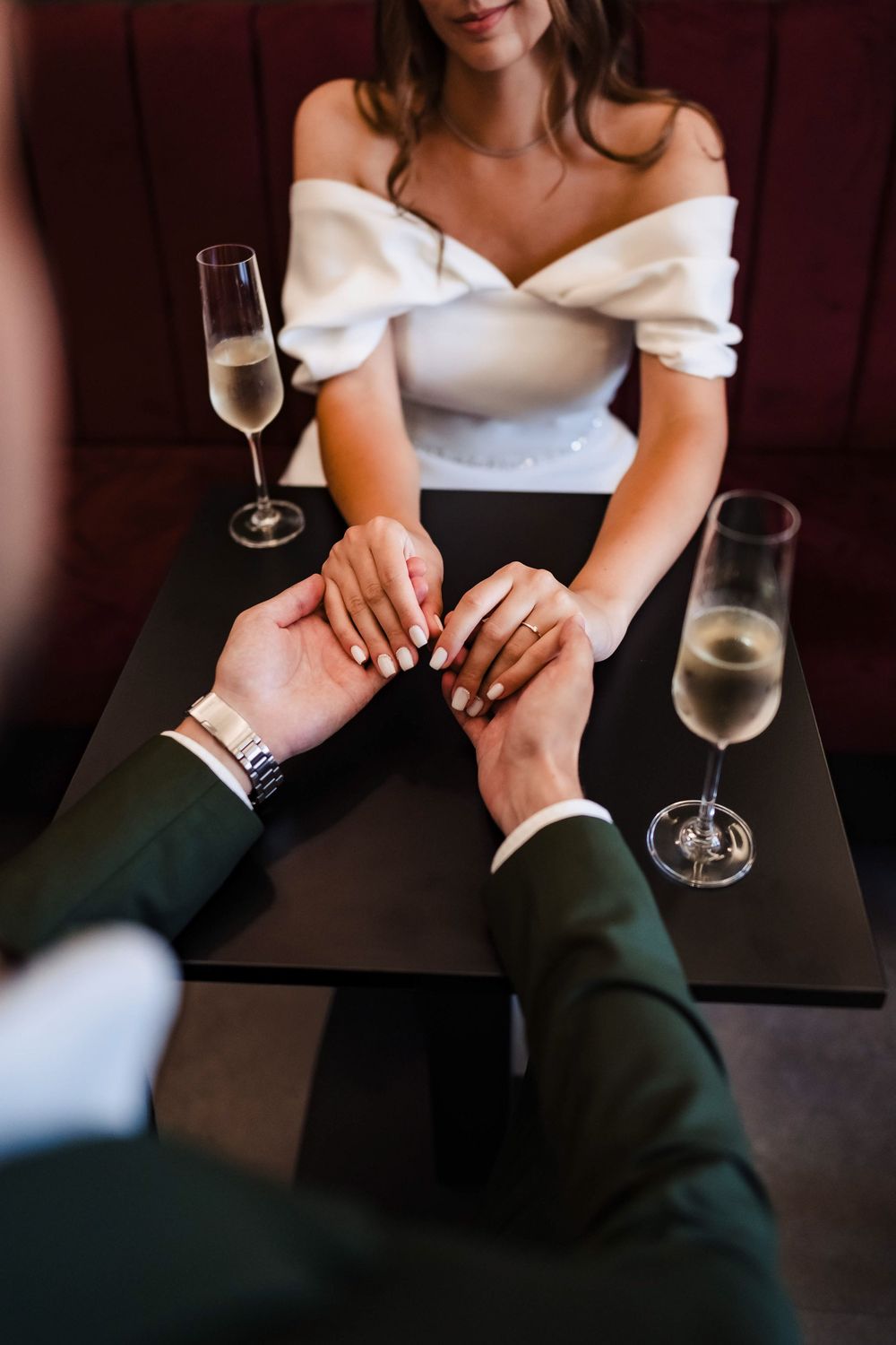Couple holding hands at a romantic dinner table with champagne glasses, bride in white off-shoulder dress.