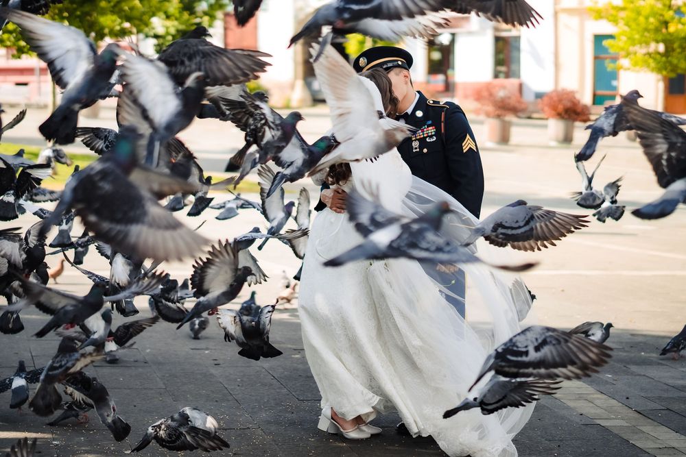 Military groom in uniform dances with bride in white dress as pigeons fly around them in an outdoor plaza.