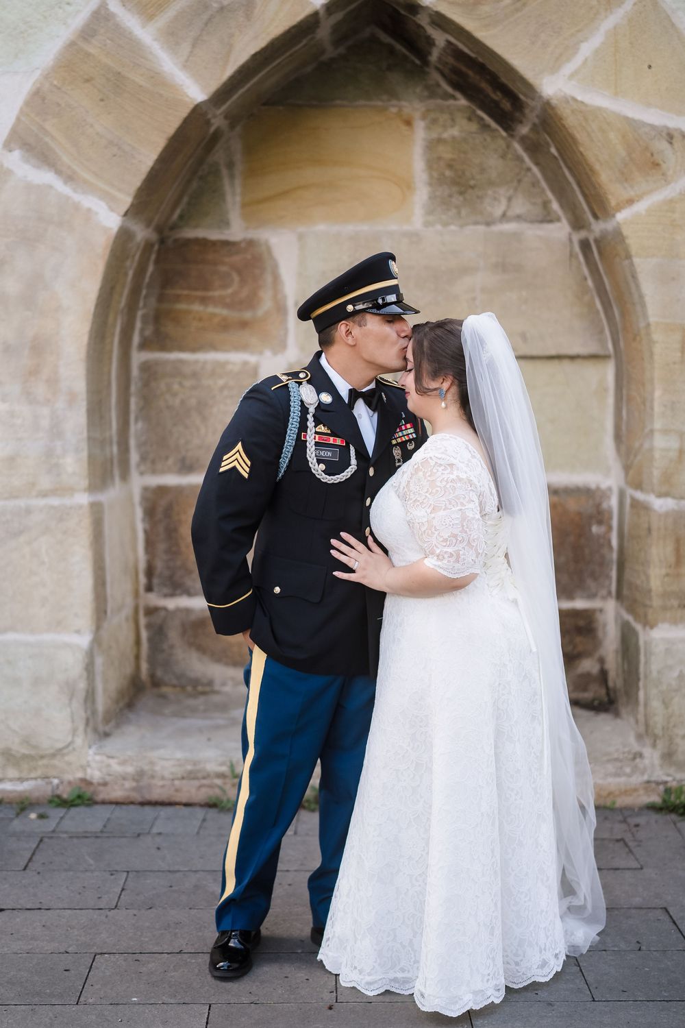 A military groom in dress uniform kisses his bride in a white lace gown and veil before a stone arch.