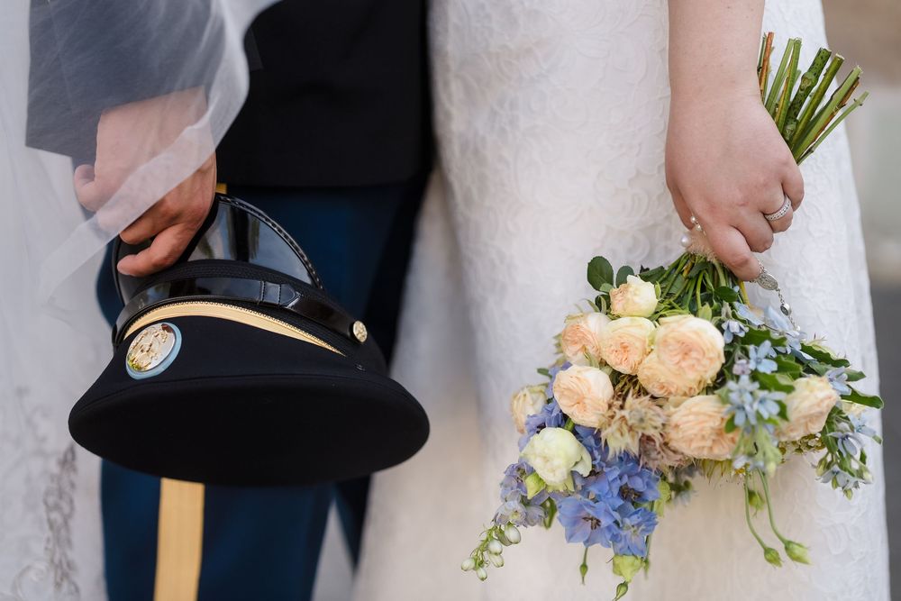 Military groom holds dress cap beside bride with pastel pink and blue wedding bouquet of roses and wildflowers.