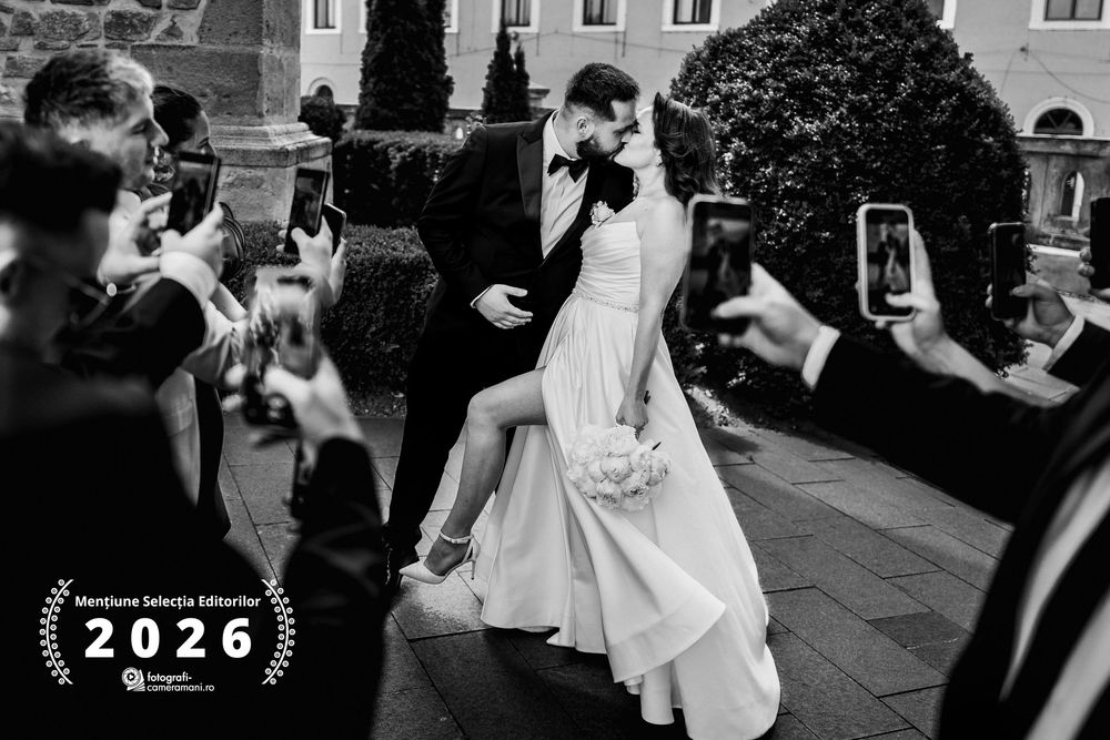 Black and white wedding photo of couple sharing romantic dip kiss while guests celebrate and photograph the moment.