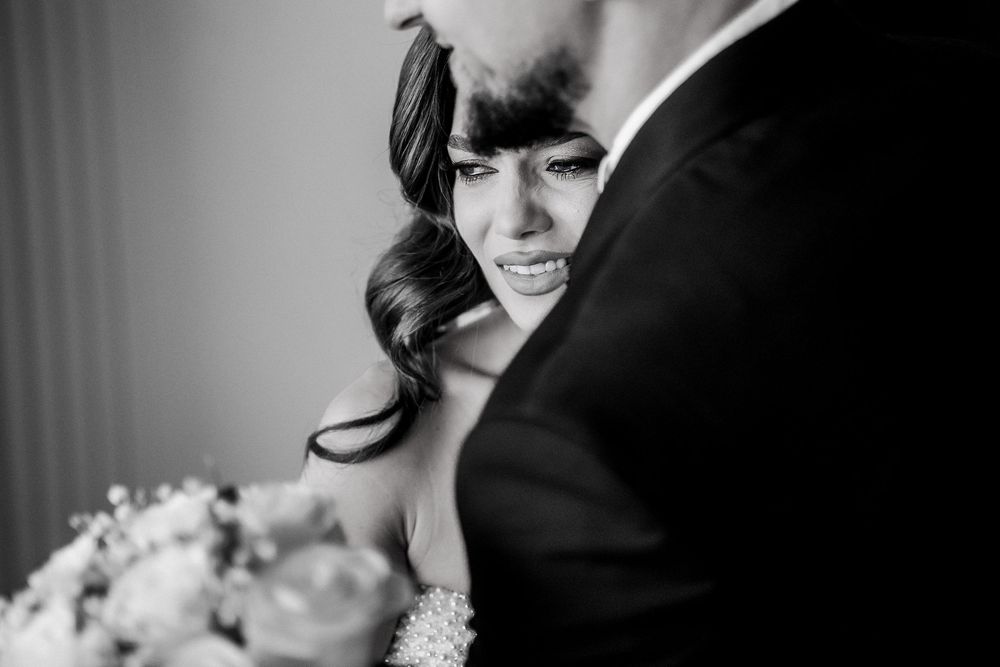 Black and white wedding photo of bride leaning against groom, holding a bouquet, with an emotional expression.