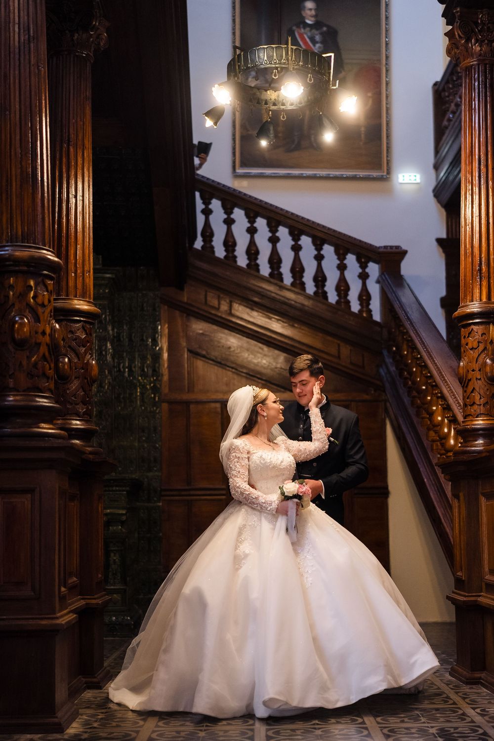 Bride in white ball gown and groom in tuxedo pose on ornate wooden staircase in elegant historic venue.