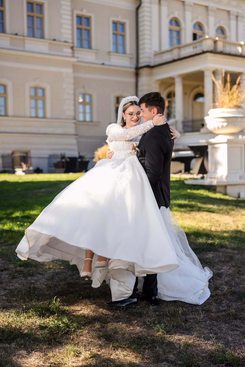 Bride in white ball gown embraces groom in black suit outdoors near elegant yellow building on sunny wedding day.