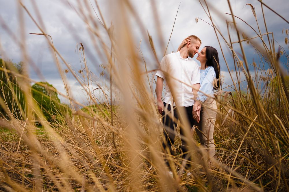 Couple sharing a romantic moment surrounded by tall golden grass in an outdoor field under a cloudy sky.