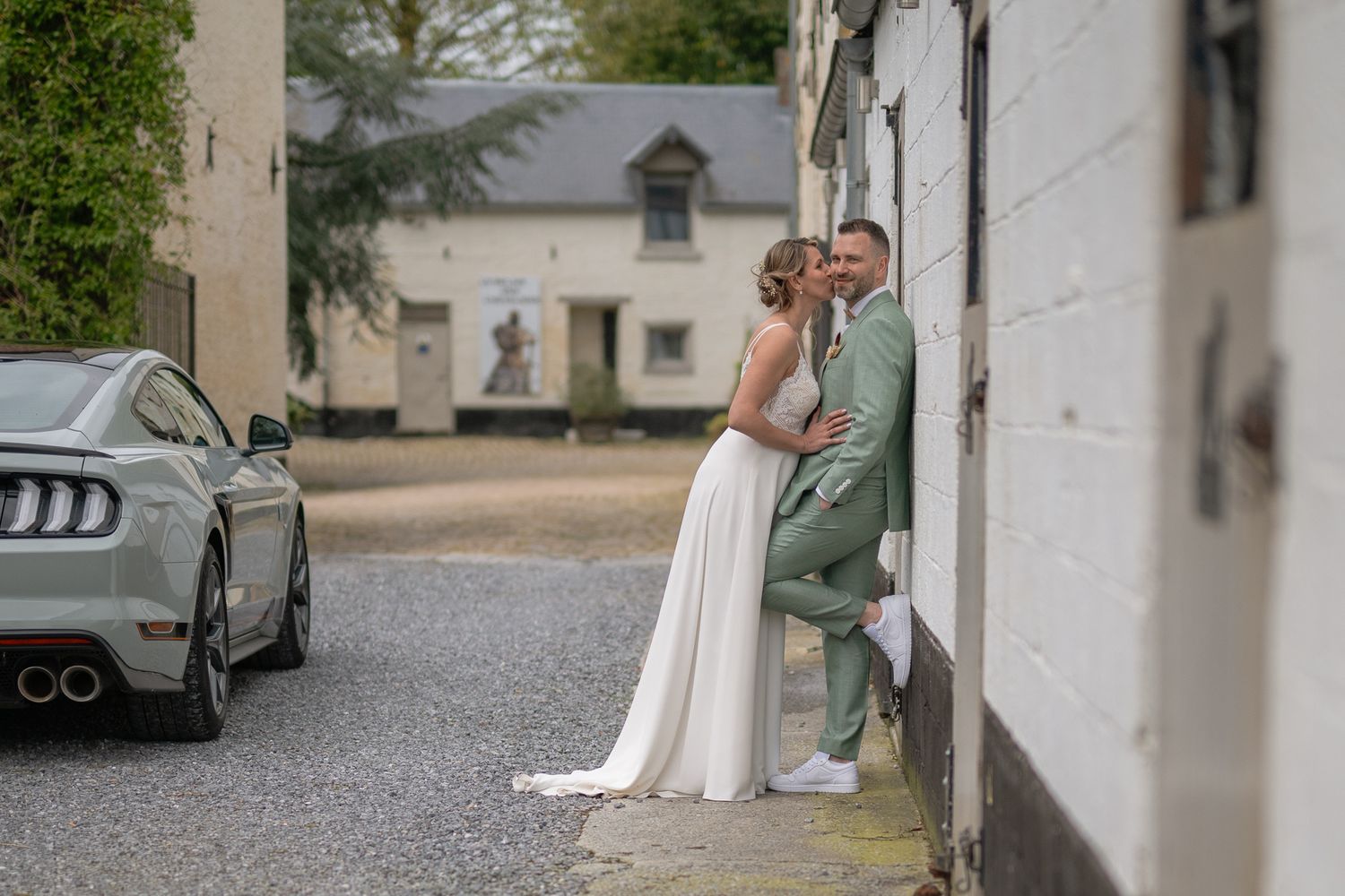 Scène de mariage élégante avec les mariés posant devant un bâtiment blanc et une voiture de luxe.