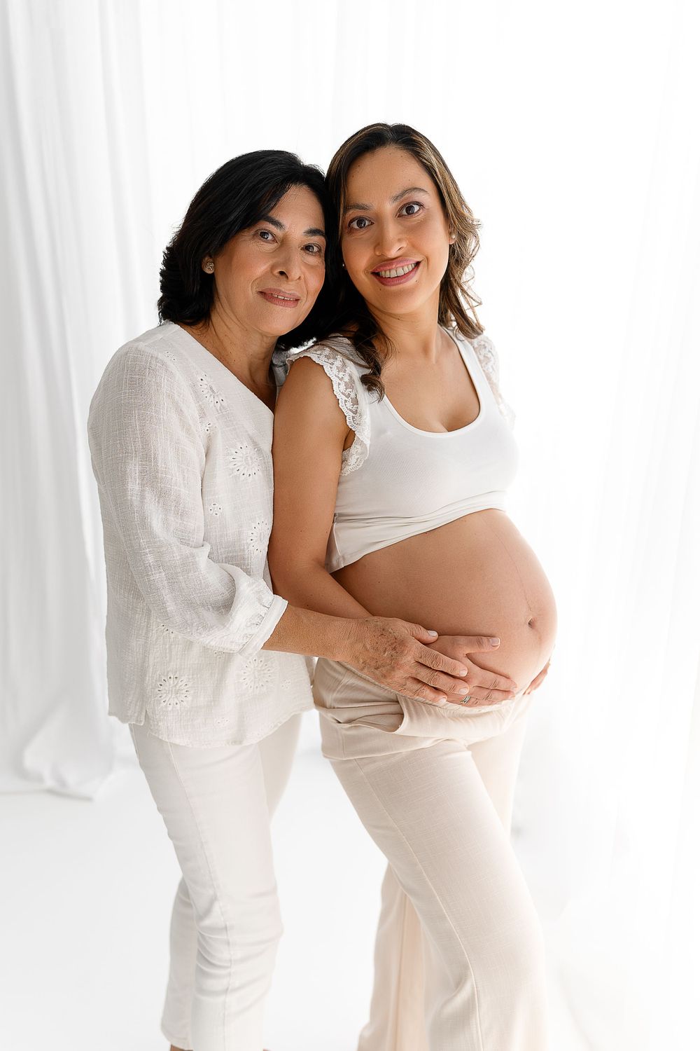 Maternity portrait of two women in white clothing embracing against bright background.