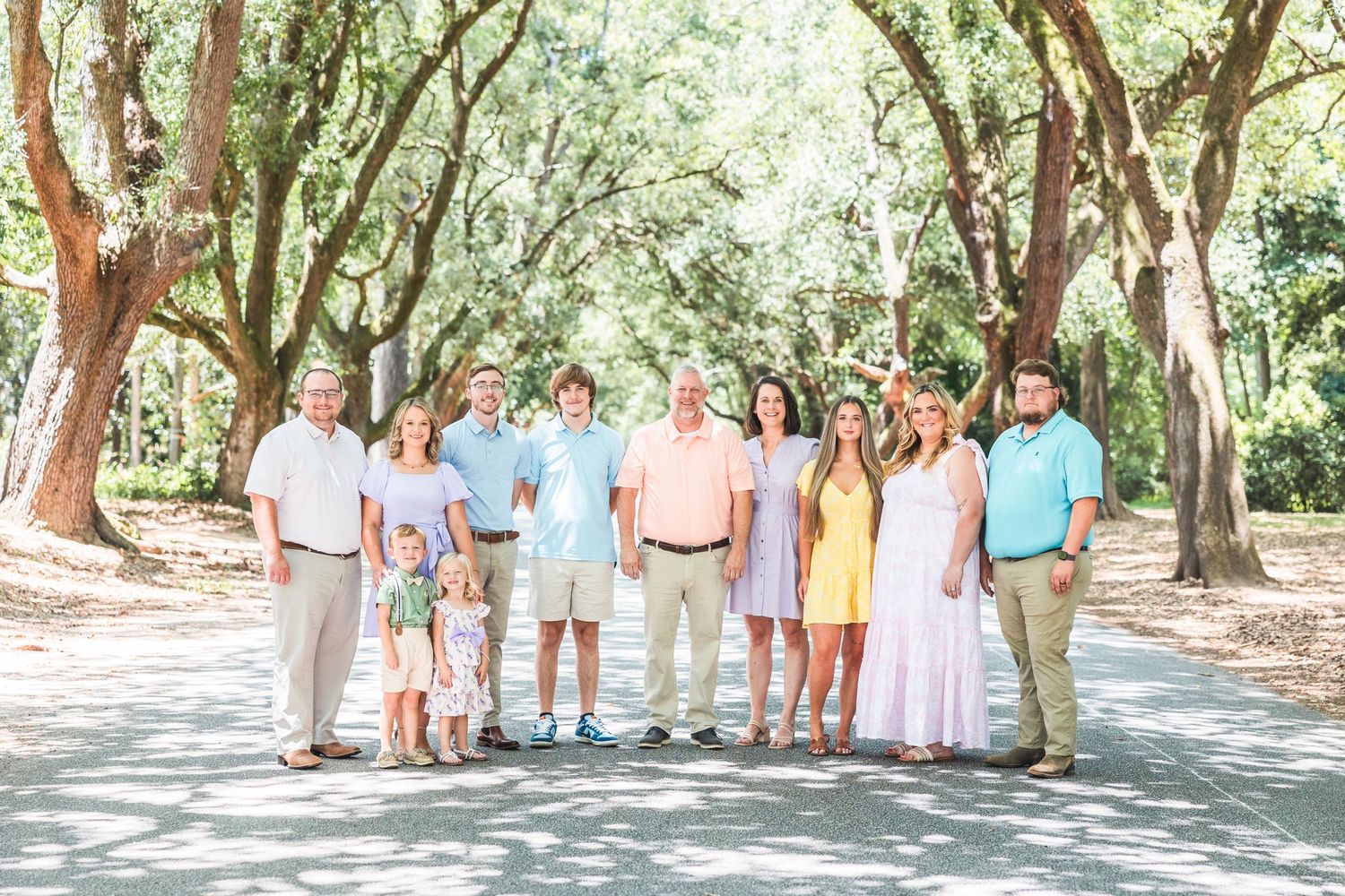 Large group photo of people in pastel clothing gathered under oak trees.