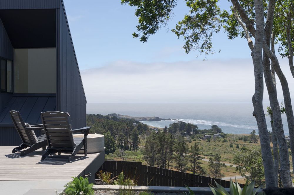 Two Adirondack chairs on a wooden deck overlooking a scenic coastal landscape with trees and ocean views.
