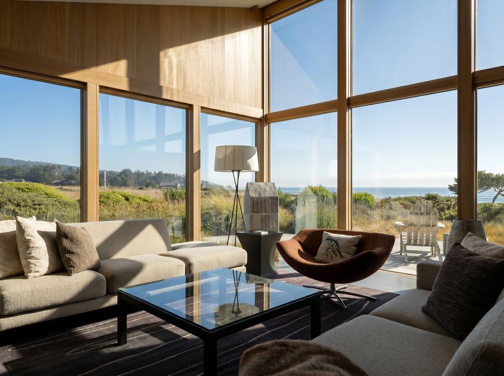 Modern living room with floor-to-ceiling windows overlooking coastal landscape, featuring gray sofas and a sleek glass coffee table.