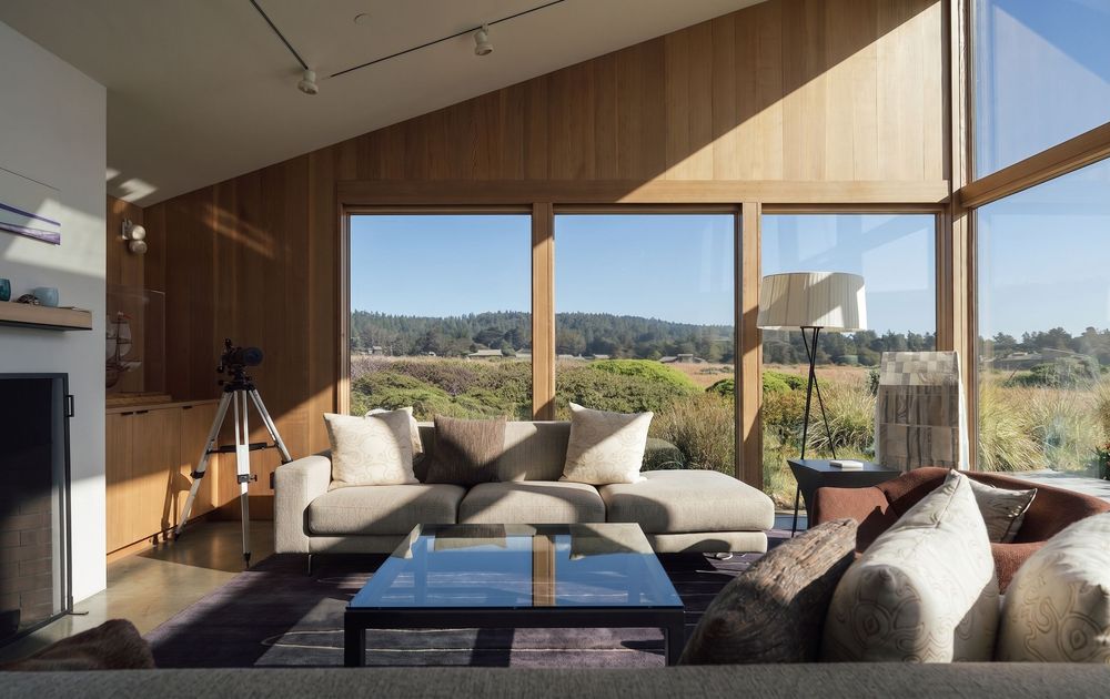 Modern living room with floor-to-ceiling windows, neutral sofas, glass coffee table, and panoramic views of rolling hills.