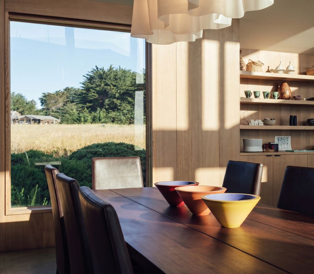 Modern dining room with wooden table, colorful bowls, and large window overlooking a sunny meadow with green trees.