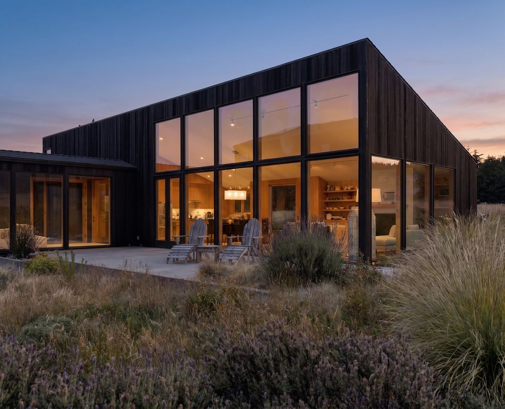 Modern black-clad home with floor-to-ceiling windows glowing warmly at dusk, surrounded by native grasses.