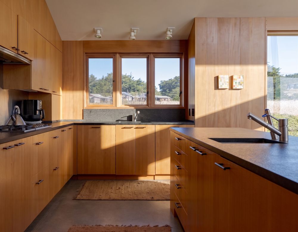 Modern kitchen with warm wood cabinetry, dark countertops, stainless sink, and natural light from large windows.
