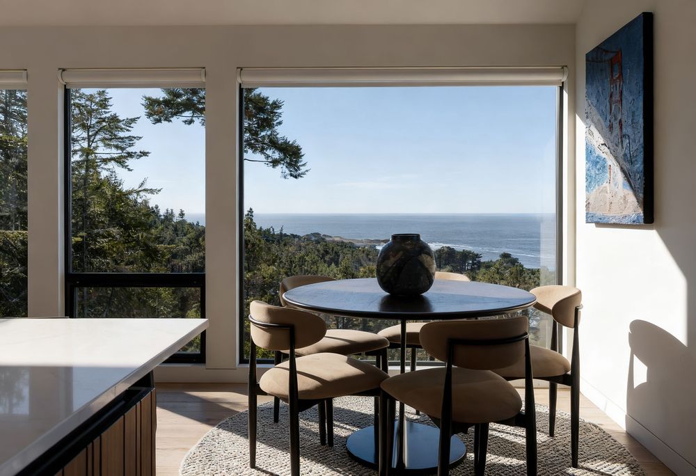 Modern dining area with round table and chairs overlooking stunning ocean and forest views through large windows.