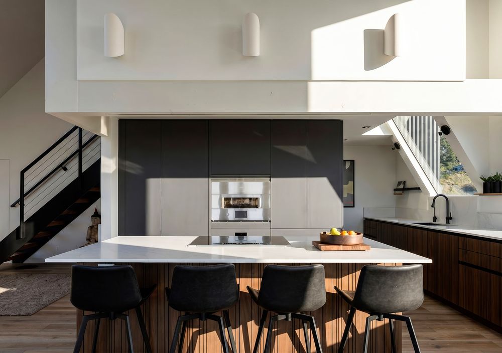 Modern kitchen with white island, black bar stools, dark cabinetry, hardwood floors, and pendant lights in an open-plan home.