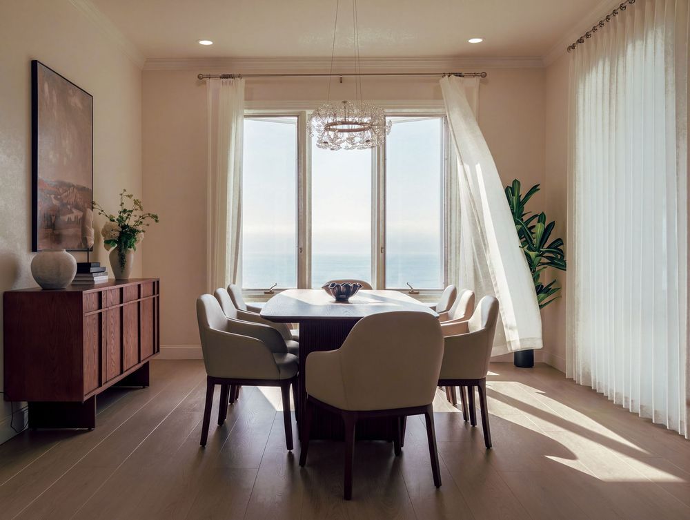 Elegant dining room with round table, beige chairs, chandelier, and ocean view through sheer curtained windows.