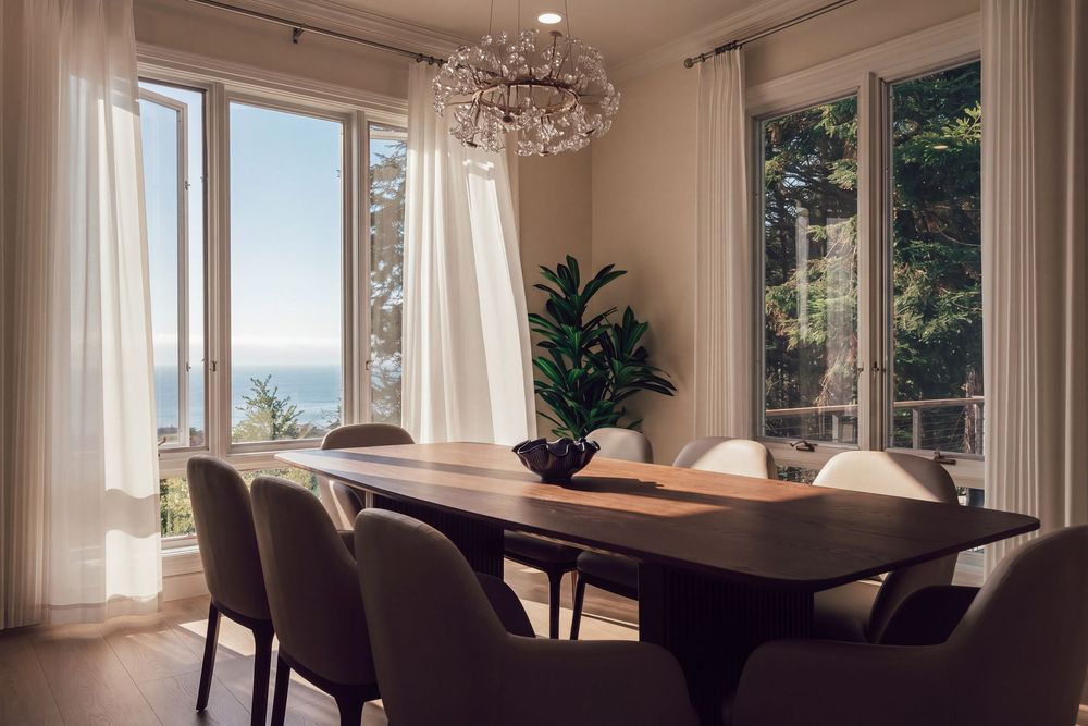 Elegant dining room with wooden table, gray chairs, crystal chandelier, and floor-to-ceiling windows overlooking ocean views.