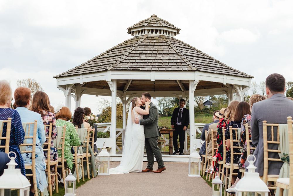 Wedding ceremony in a white gazebo surrounded by guests in garden chairs.