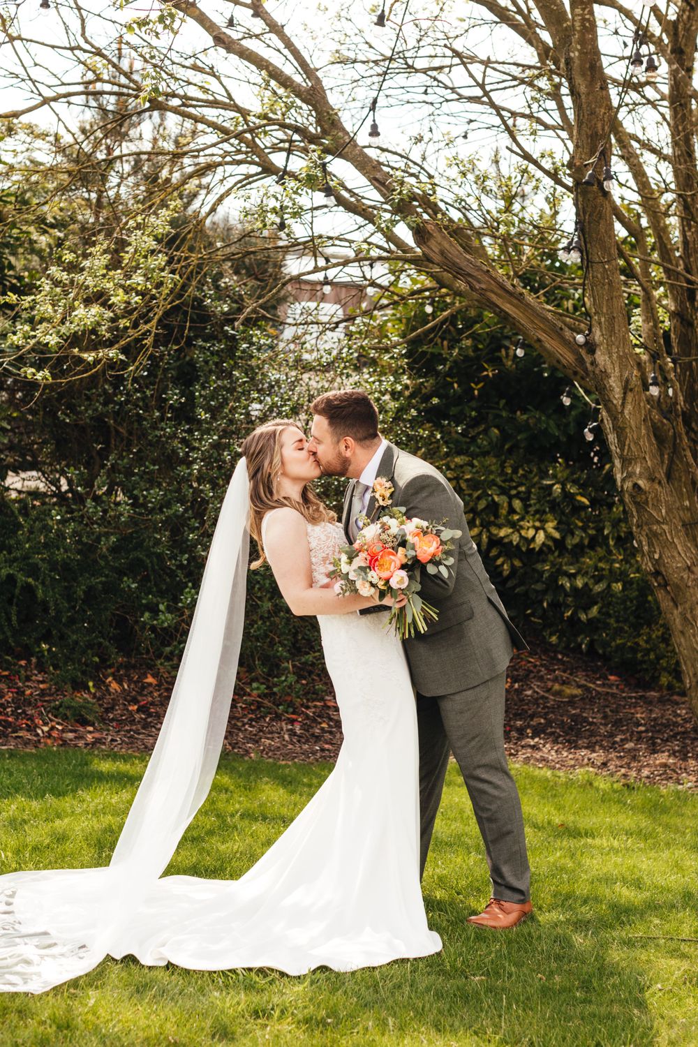 A romantic outdoor wedding photo showing a couple embracing on a green lawn with trees in the background.