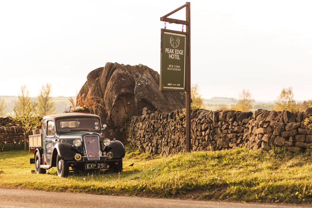 Vintage car parked beside a rustic stone wall and wooden sign at sunset.