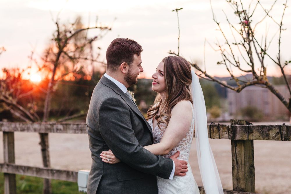Beautiful sunset wedding portrait with bride and groom embracing near wooden fence.