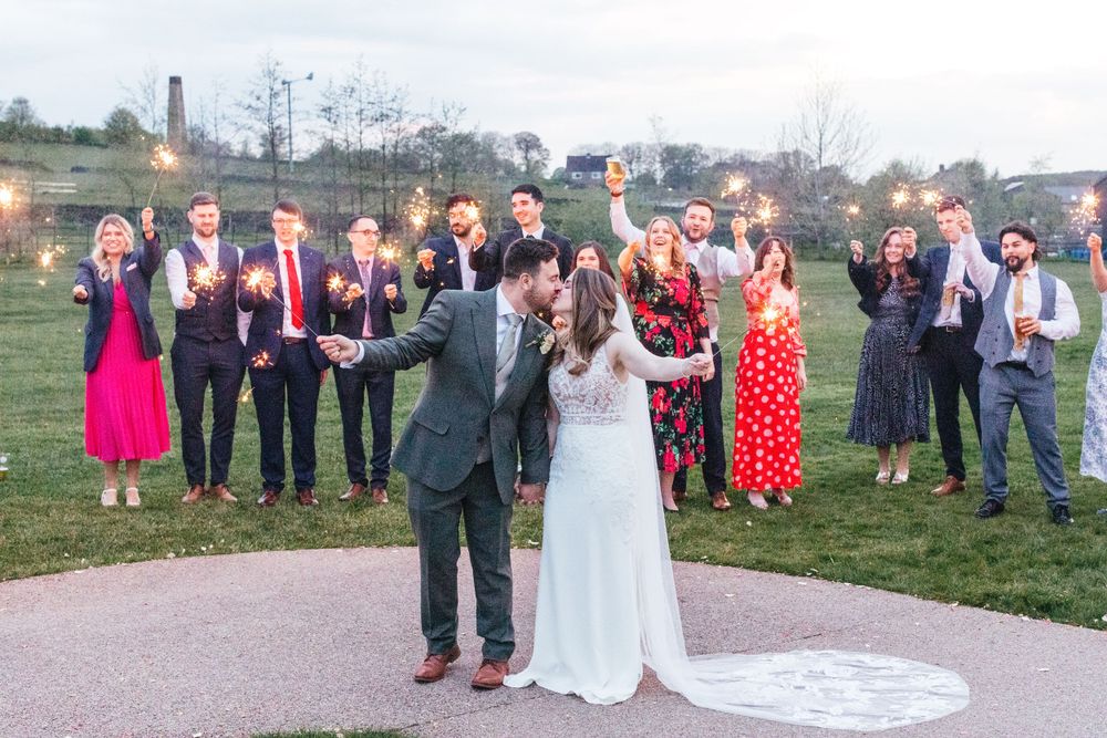 Wedding celebration with wedding party cheering in background while couple kisses.