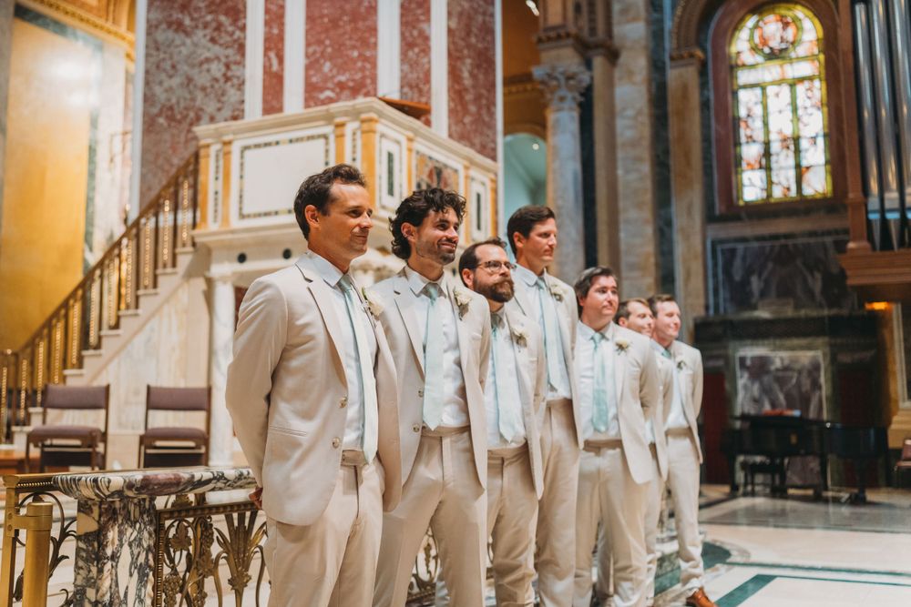Groomsmen in light colored suits standing in a row inside an ornate church.