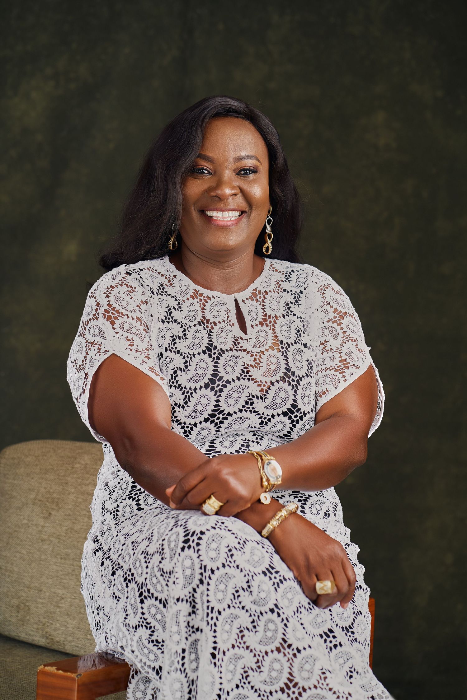 Portrait in white lace dress with jewelry sitting against dark green background.