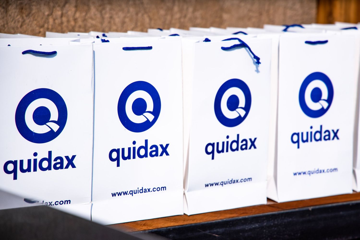 Row of white shopping bags with blue Quidax logo and branding displayed.