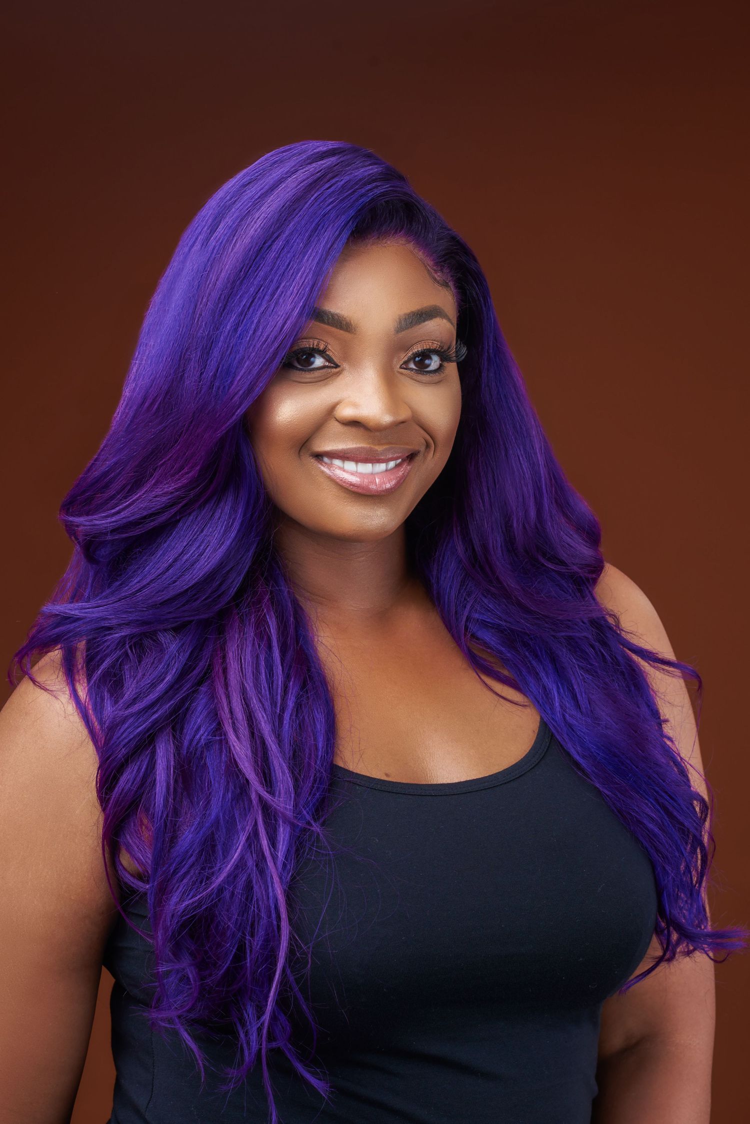 Portrait of person with vibrant purple wavy hair wearing black top against brown background.