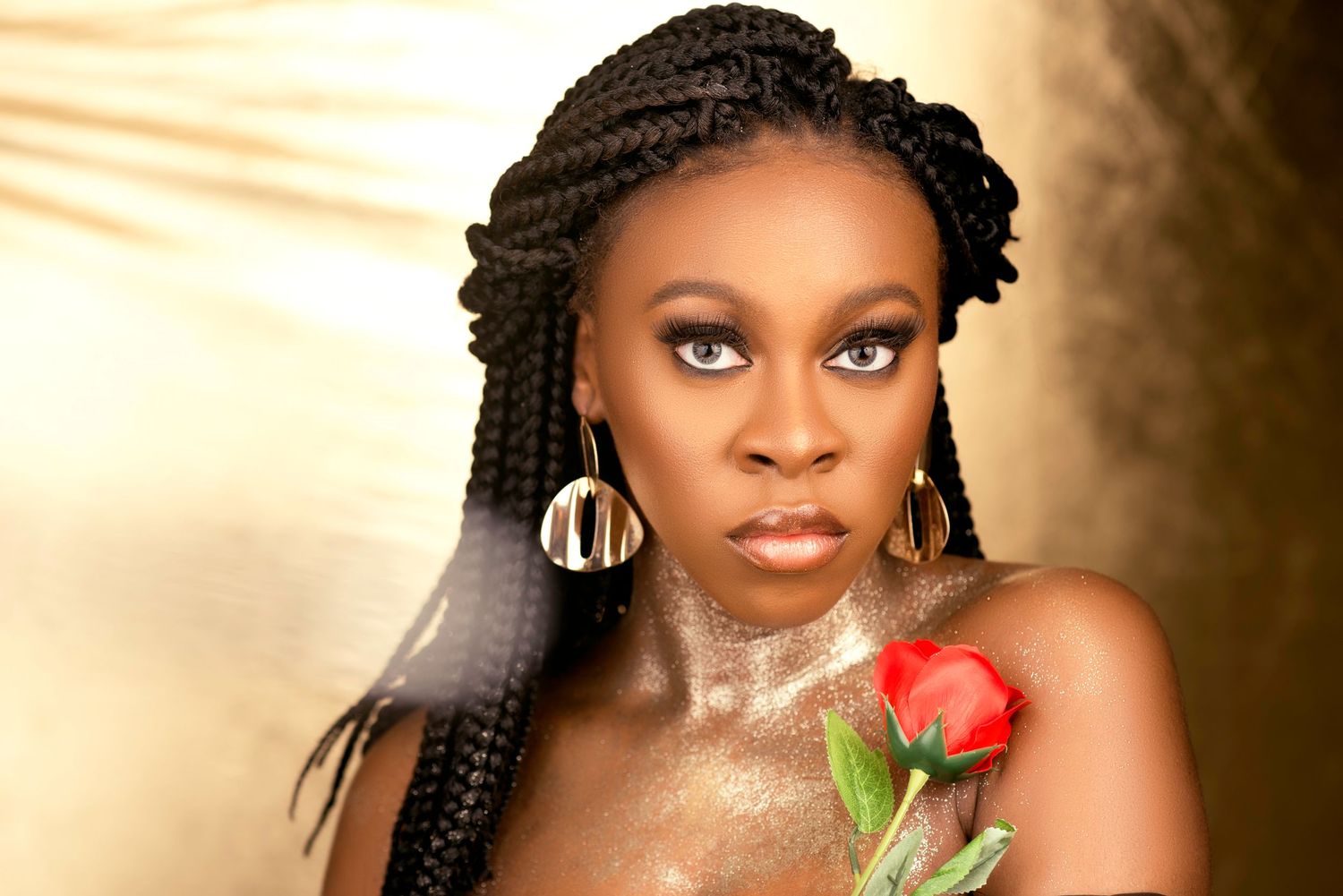 Glamorous portrait with braided hair, dangling earrings, and red rose against golden background.