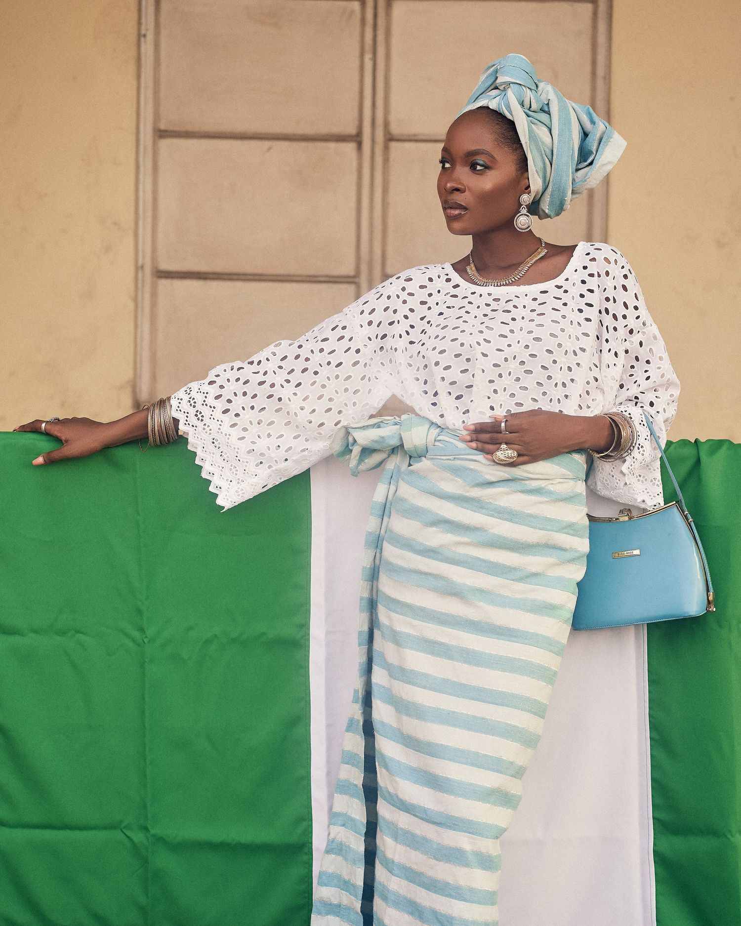 A stylish outfit featuring a white crochet top, striped wrap skirt, light blue headwrap, and matching handbag against a green backdrop.
