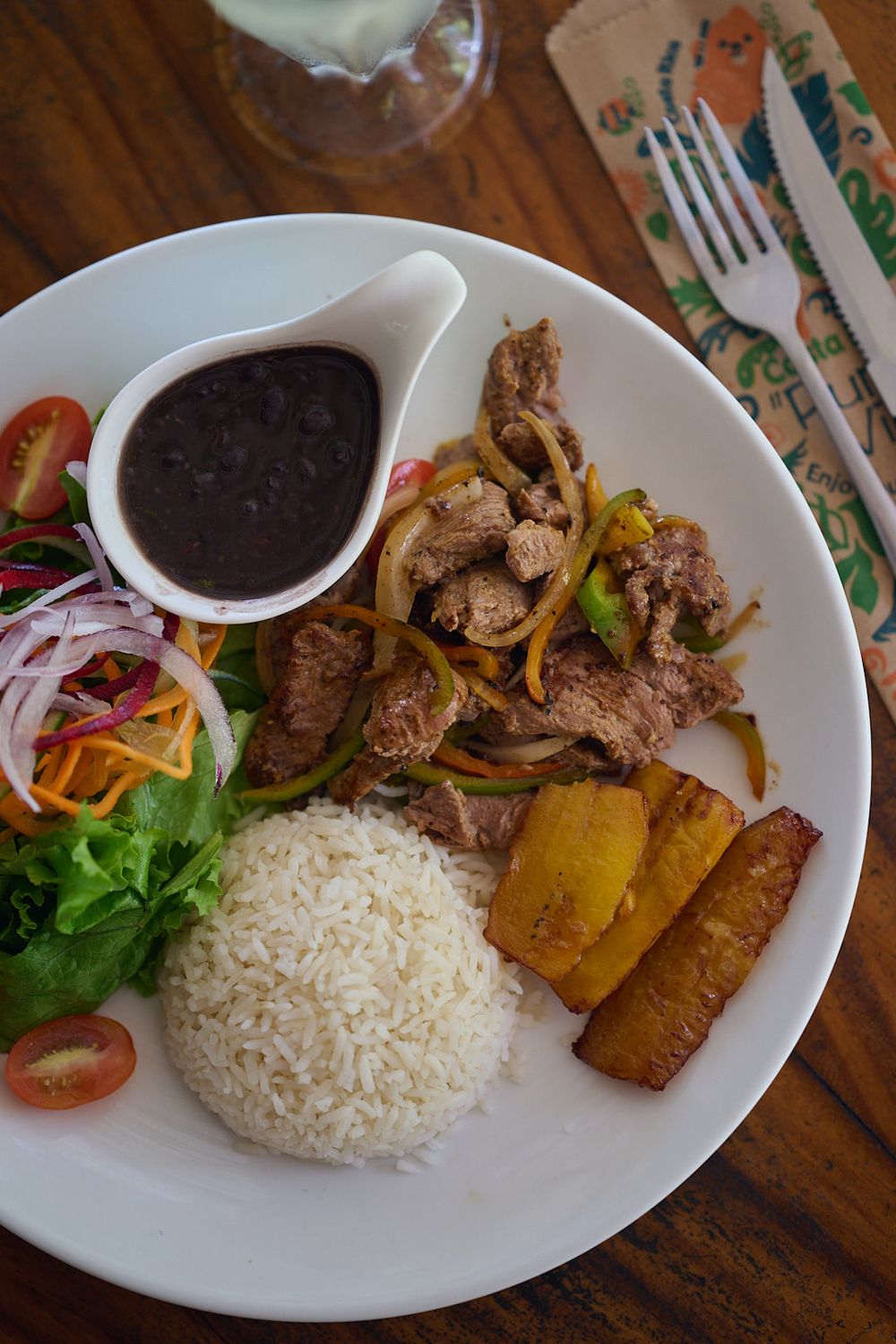 Peruvian plate with lomo saltado, white rice, fried plantains, black sauce and fresh salad on a wooden table.