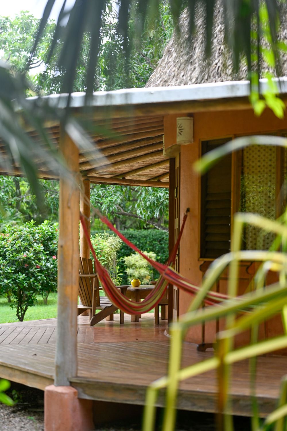 A cozy wooden deck porch with a red hammock hanging under a rustic roof surrounded by lush tropical greenery.