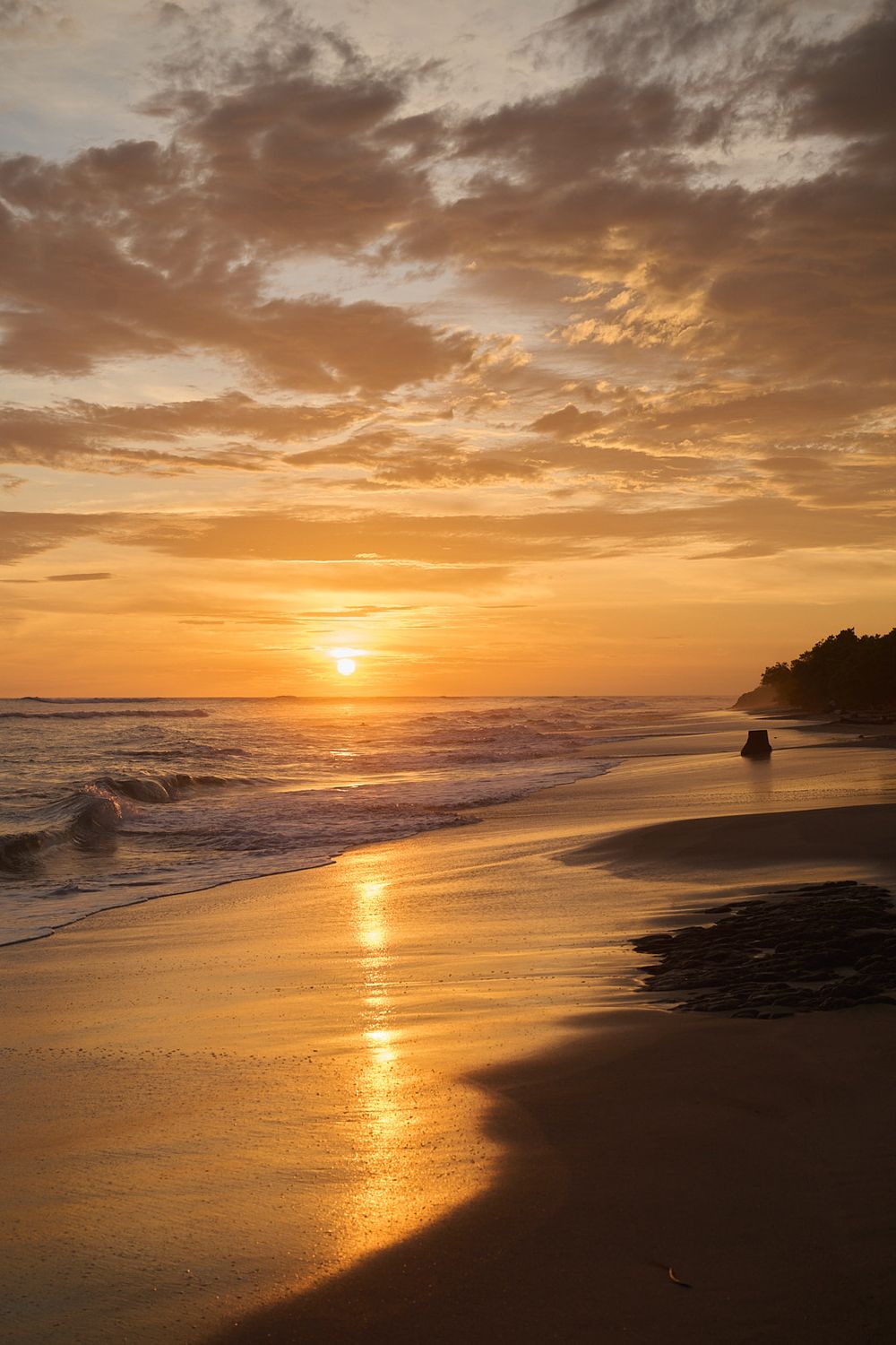 Stunning ocean sunset with golden light reflecting off waves breaking on sandy beach as dramatic clouds fill orange sky.