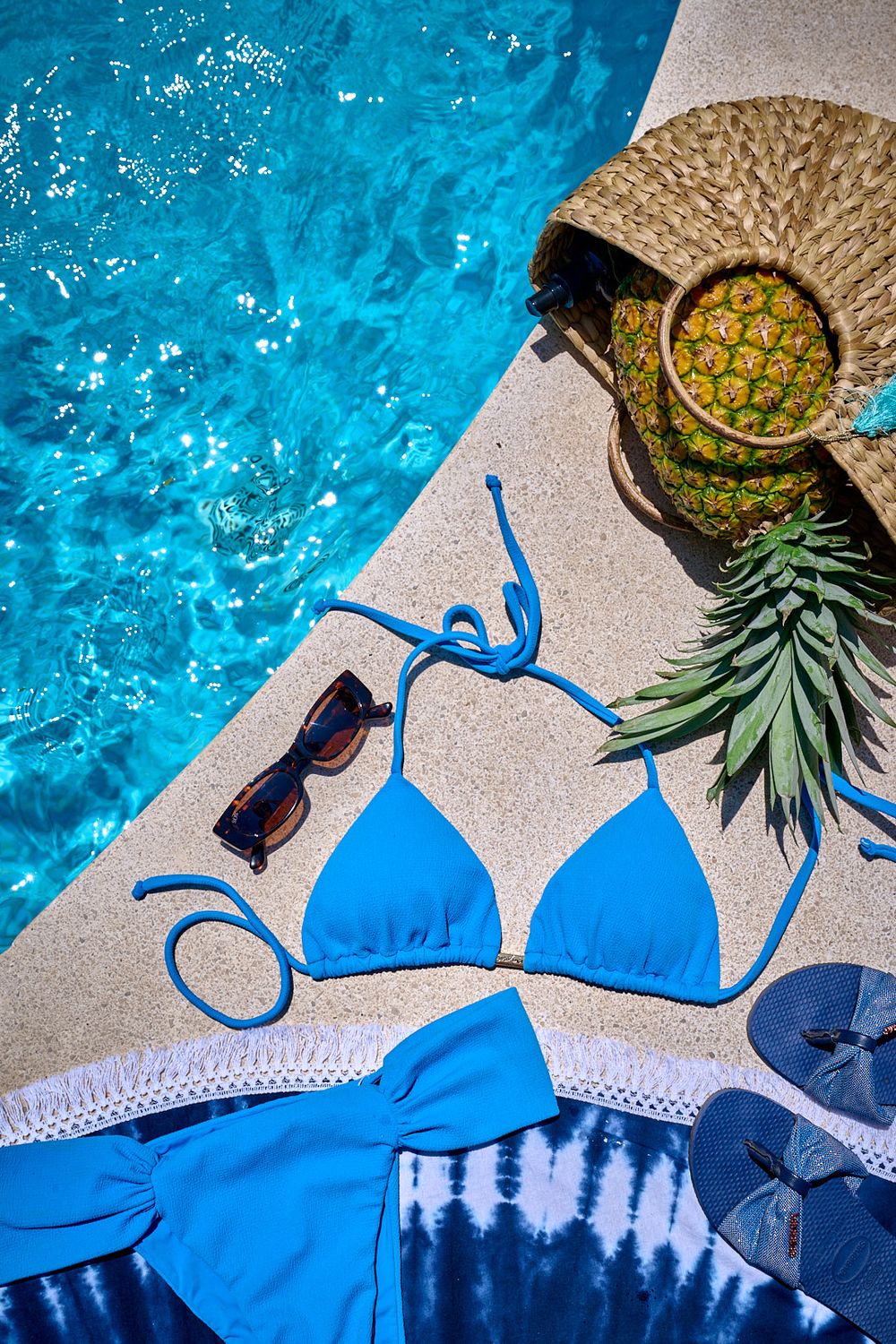 Blue bikini, sunglasses, and fresh pineapple arranged by a sparkling pool for a summer vacation lifestyle scene.