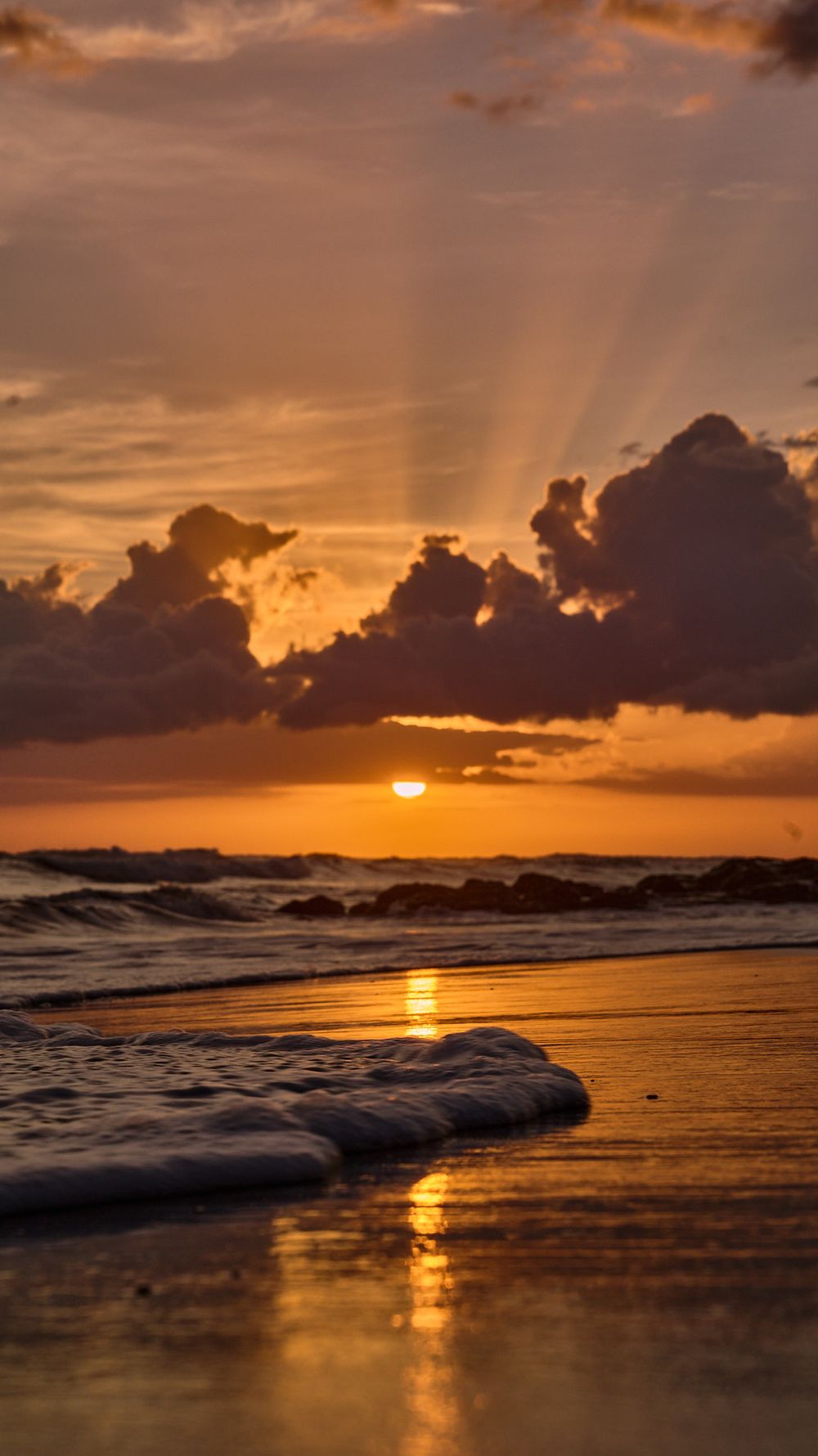 Dramatic ocean sunset with golden sun rays piercing through dark clouds and reflecting on wet sandy beach.