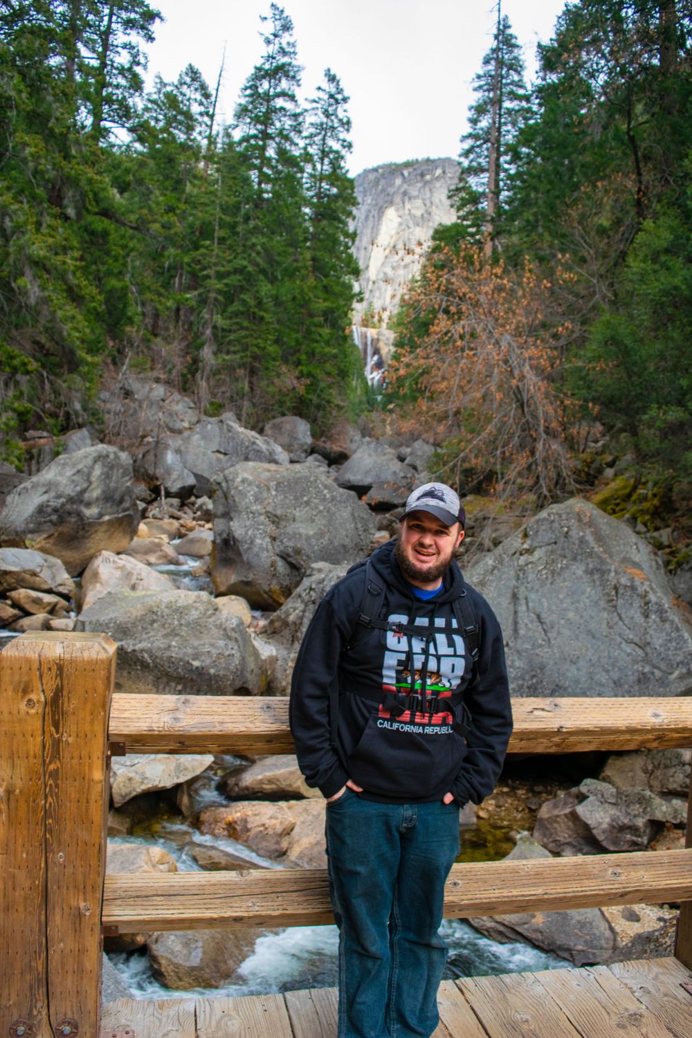 Visitor stands on wooden bridge over rushing stream with rocky cliff face and forest in background.