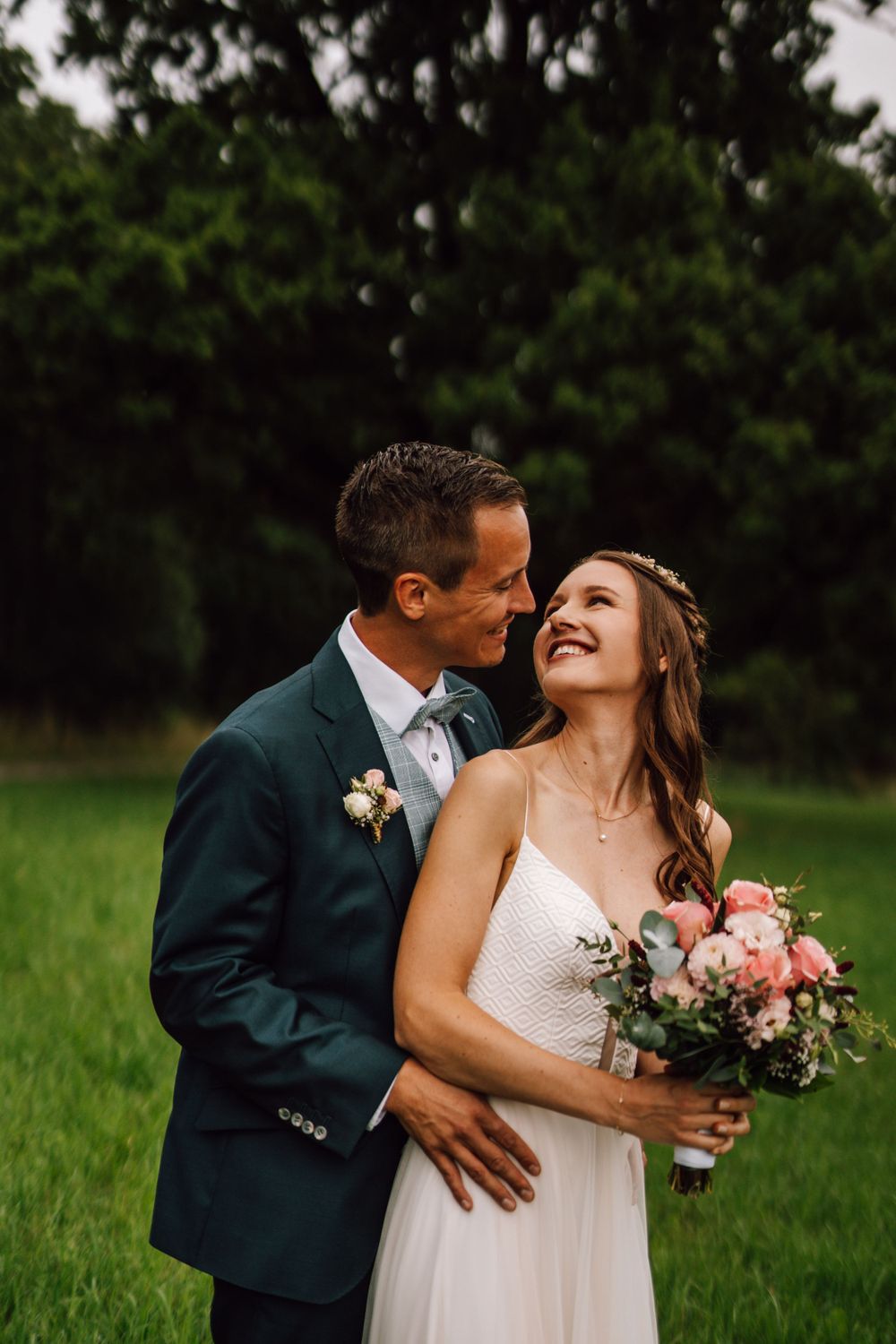 A bride holds a pink bouquet while sharing a tender moment with her groom in a grassy field.
