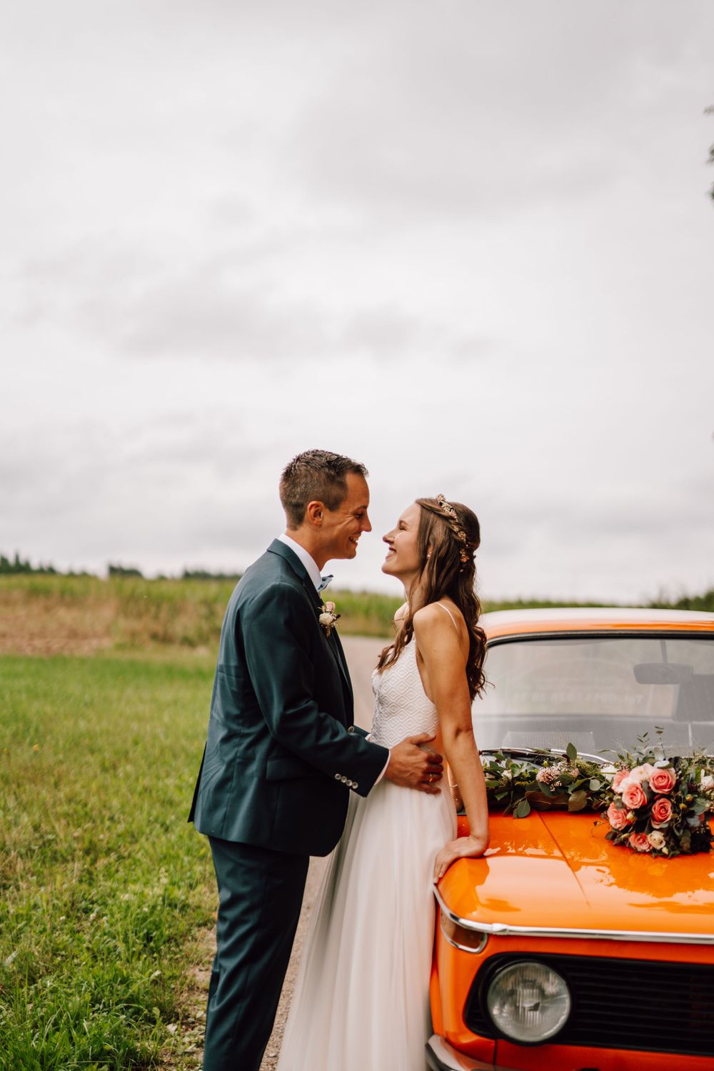 Newlyweds share romantic moments near a vintage orange car in a rustic field setting.