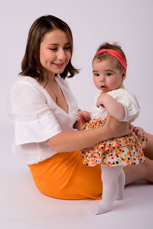 Mother in white blouse and orange skirt holding baby in floral outfit against light background.