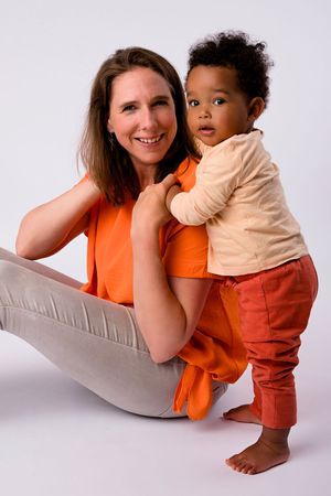 Mother in orange shirt sitting with toddler in beige top against studio background.