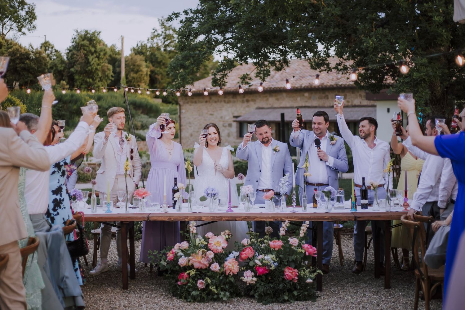 Wedding party celebrates at a long reception table decorated with flowers and string lights in an outdoor garden setting.