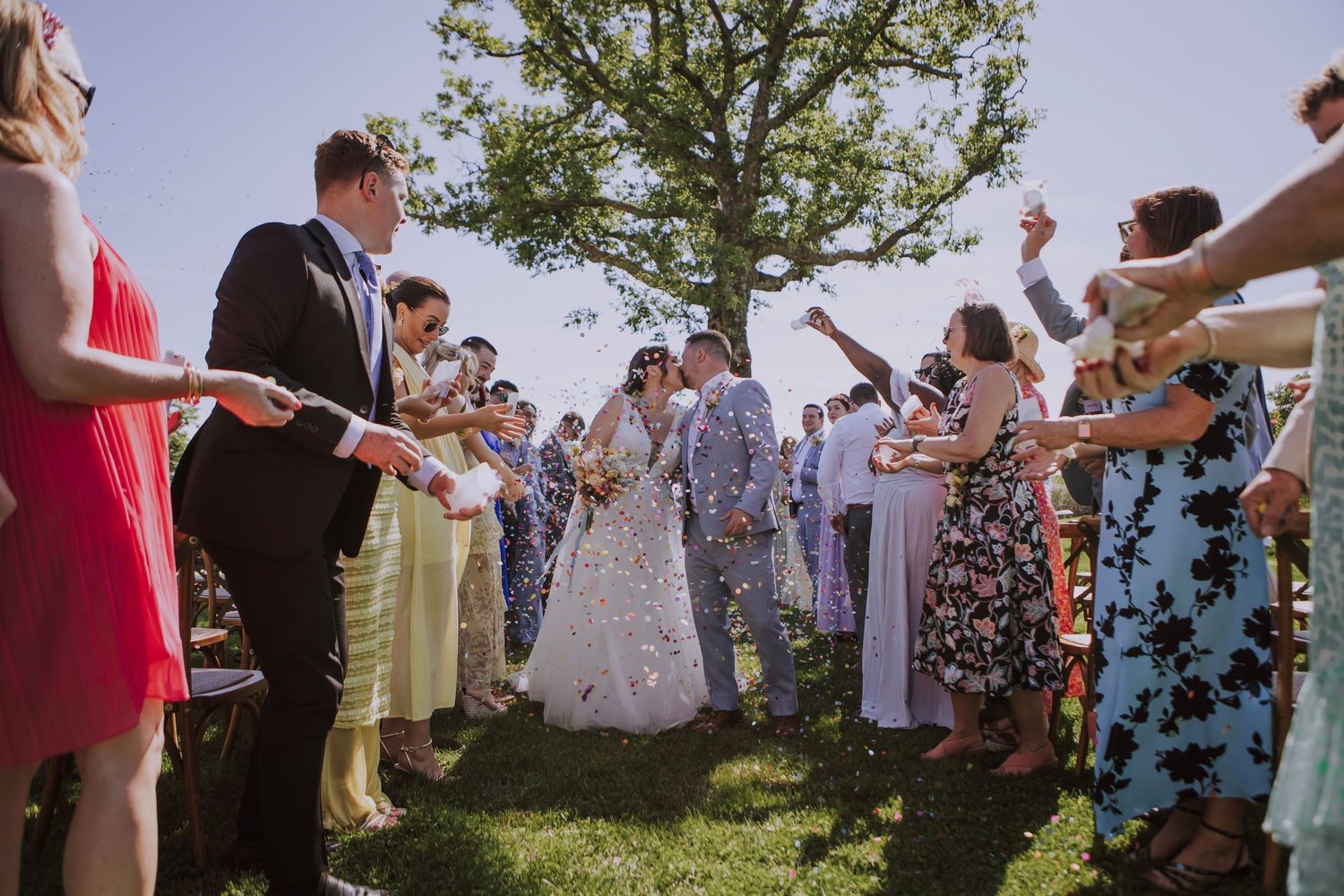 Wedding guests form a tunnel outdoors under a large tree during a celebration.