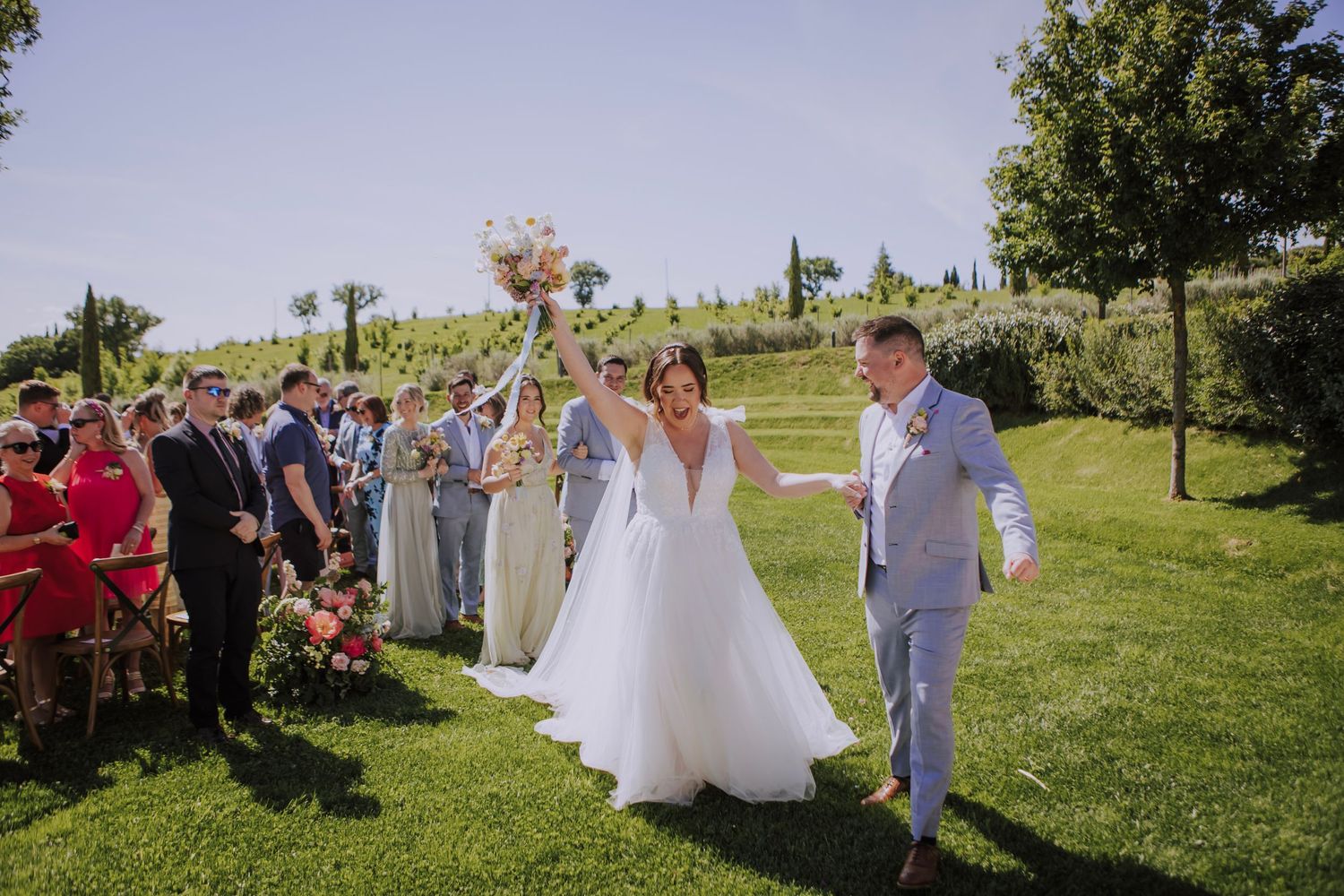 Wedding celebration on a sunny lawn with guests cheering as couple walks down outdoor aisle.