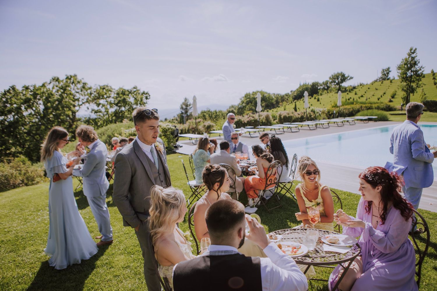 Outdoor wedding reception with guests dining at tables on a grassy hillside.