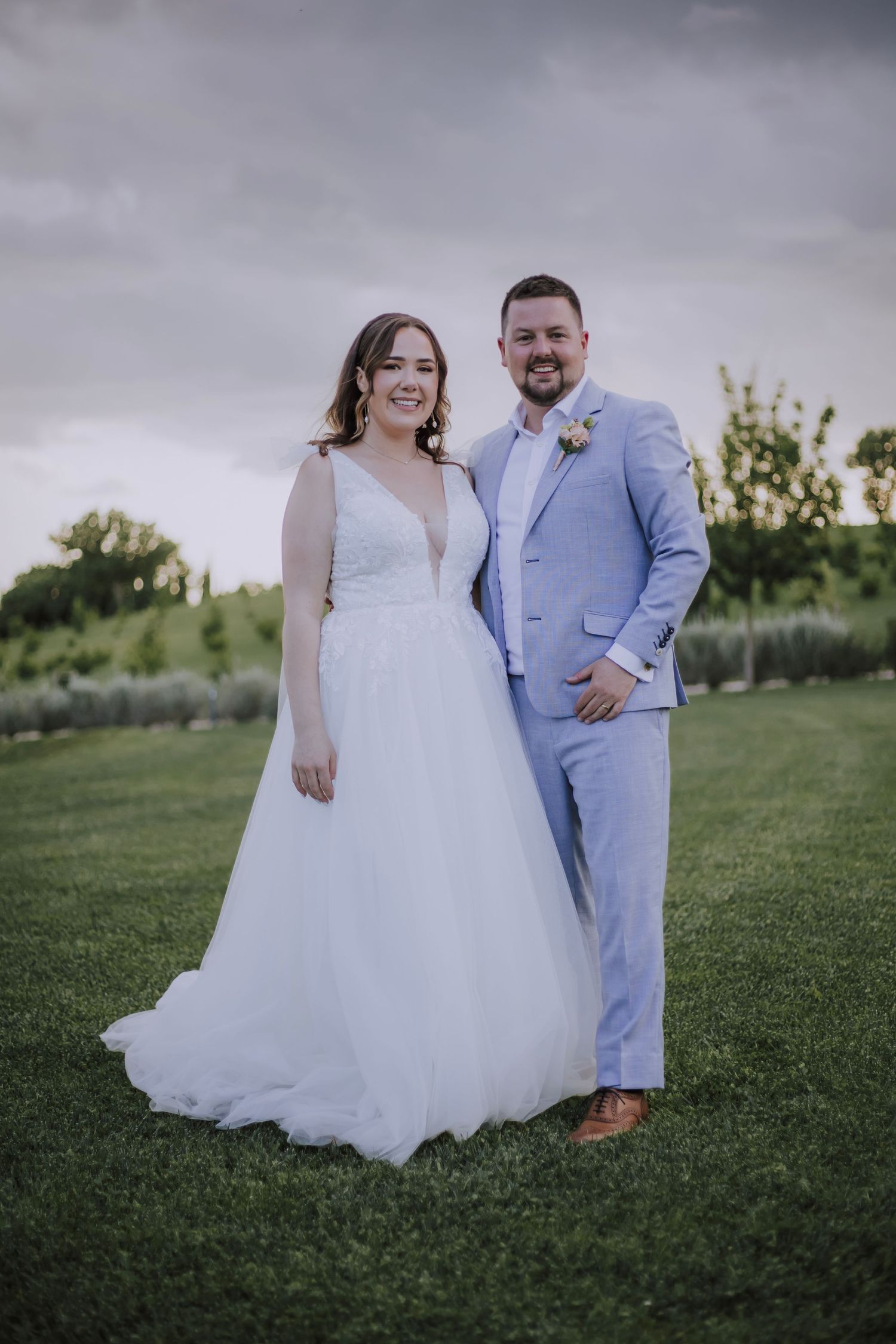 Wedding couple standing on grass with cloudy skies behind them.