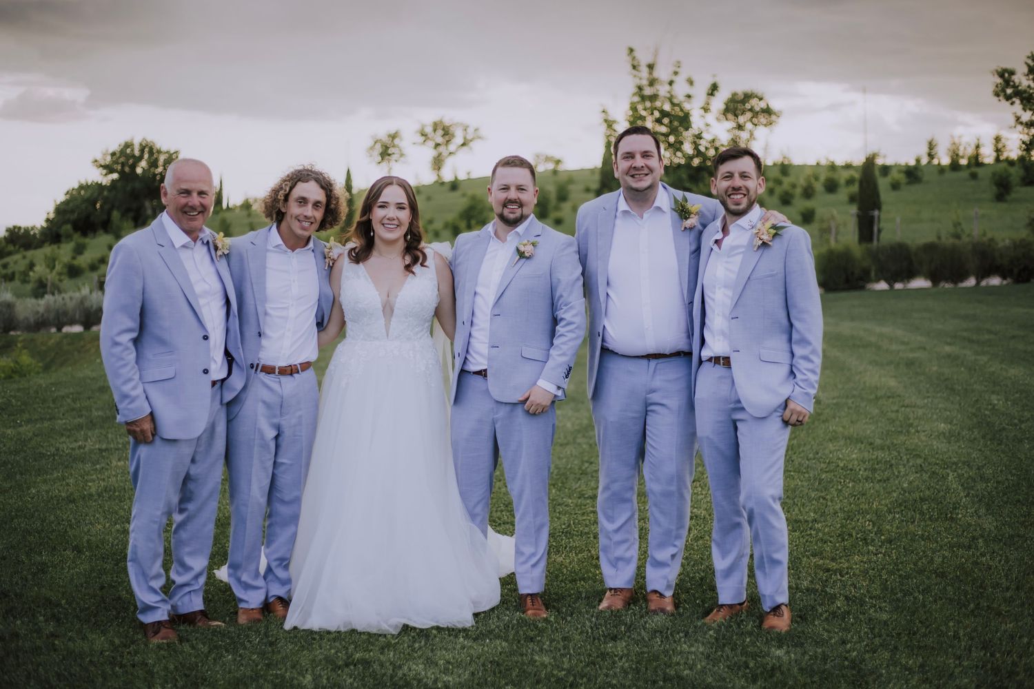 Wedding party in coordinating light blue suits posing together on grass.